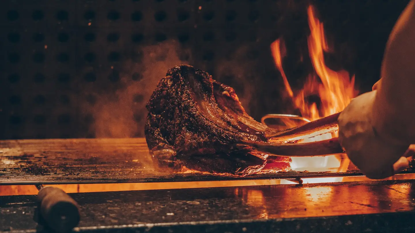 Chuletón sobre la parrilla con llamas al fondo y una mano sujetándolo con pinzas